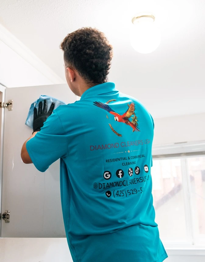 Diamond Cleaners USA professional cleaner wearing a turquoise uniform cleaning a kitchen cabinet inside a home, providing reliable residential and commercial cleaning services in Seattle.